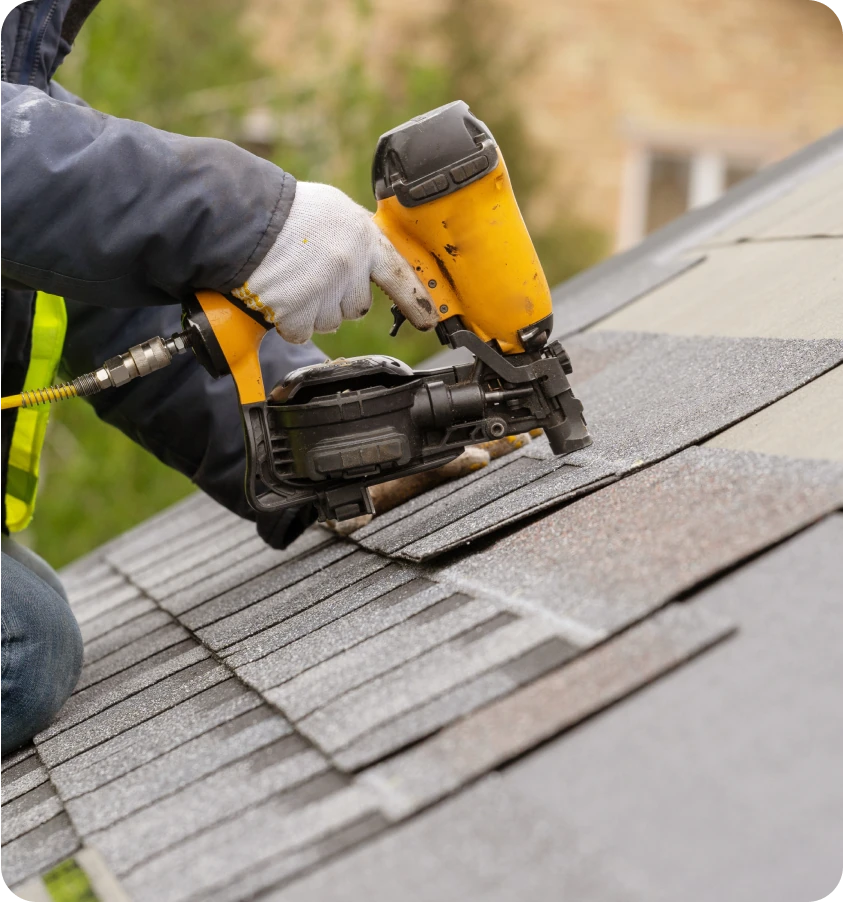 Close-up of a roofer using a yellow nail gun to install brown shingles on a home.